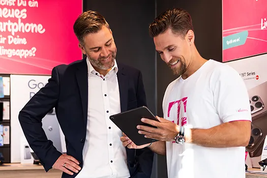 Zwei Männer im Telekom-Shop schauen auf Tablet, Beratung zu Smartphone-Tarif